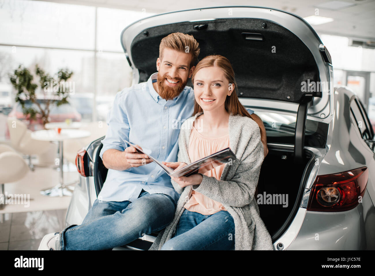 Heureux couple assis sur catalogue avec coffre de voiture au salon de l'affaire Banque D'Images
