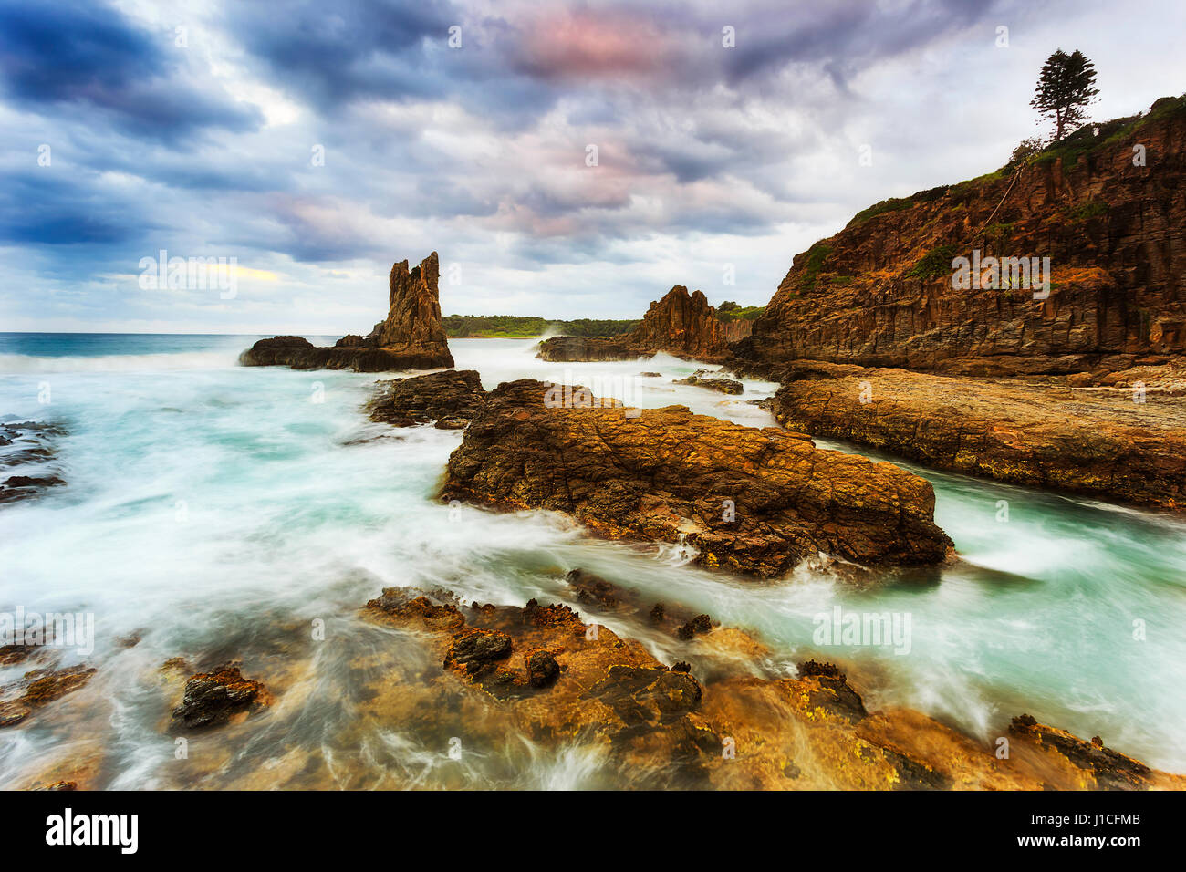 La plage de Bombo Kiama cathédrale des roches sous d'épais nuages au coucher du soleil. Les ondes brouillées sufing passez sur les blocs de grès. Banque D'Images