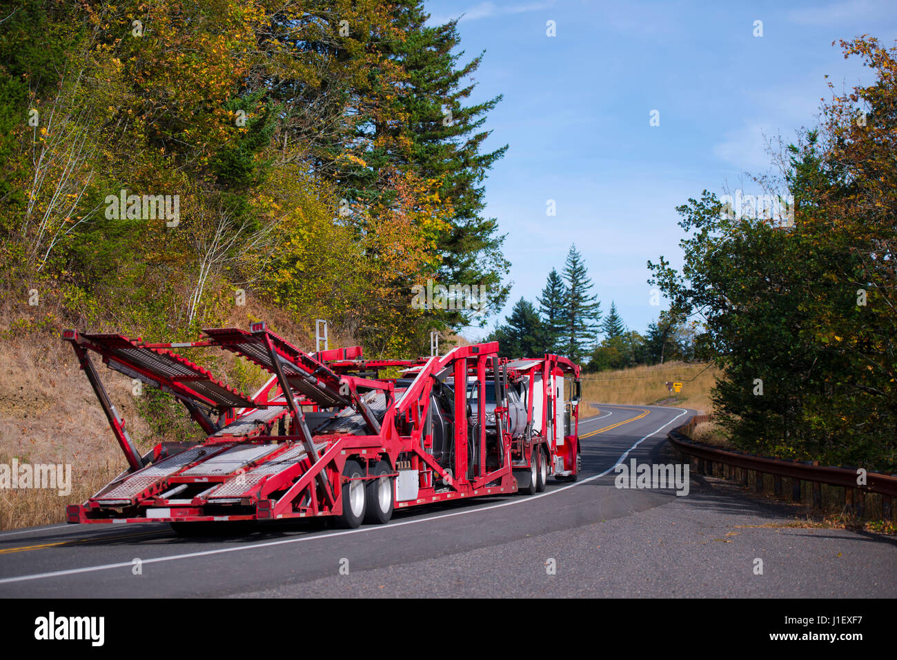 Gros camion transporteur de voiture camion semi avec remorque pour le transport spécialisé rouge plusieurs voitures se déplaçant sur une route sinueuse à travers les collines couvertes Banque D'Images