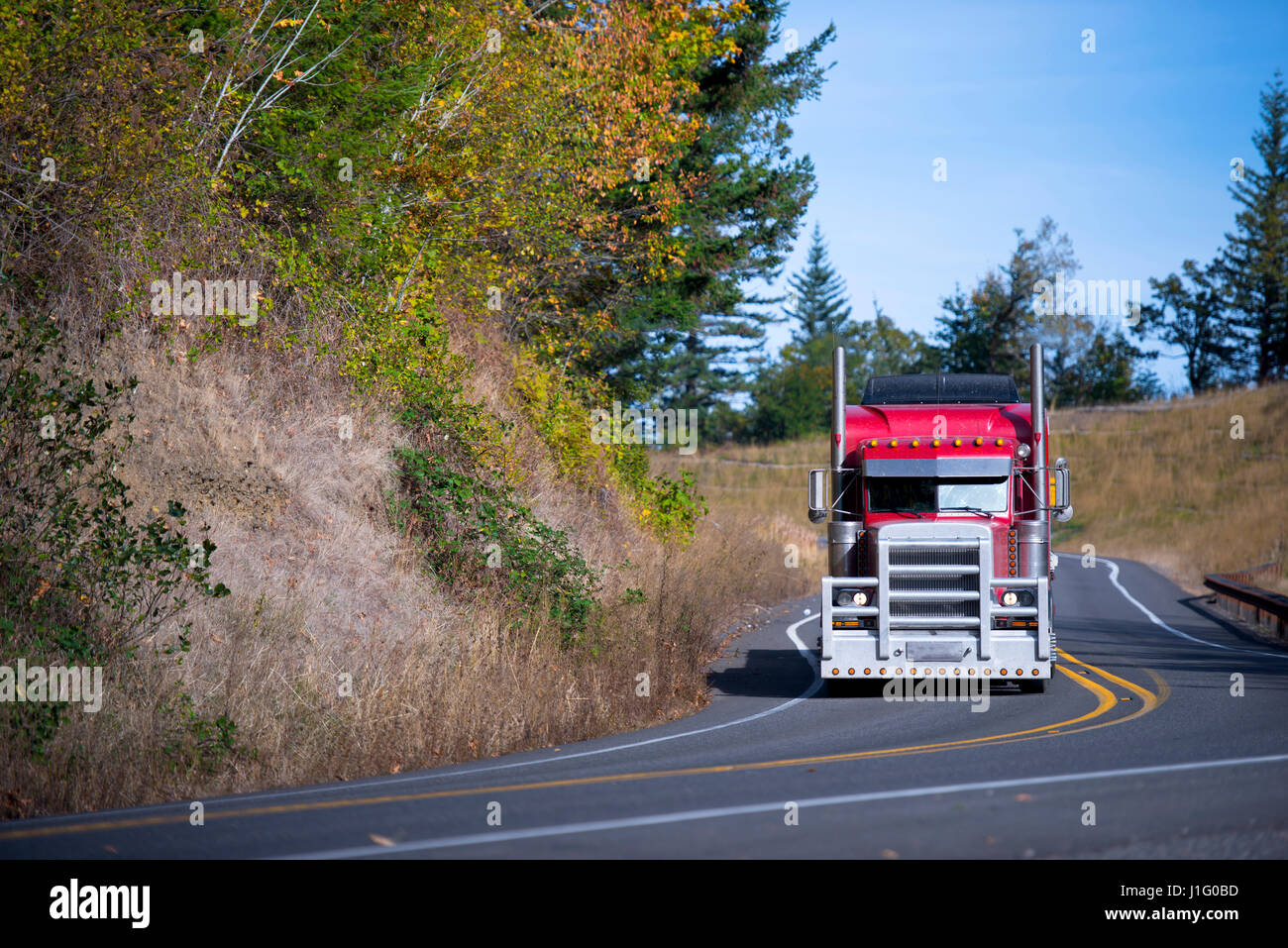 Lumineux et élégant professionnel puissant populaires rouge gros camion semi truck avec une grille de protection, semi truck va sur une route sinueuse avec les arbres d'automne. Banque D'Images