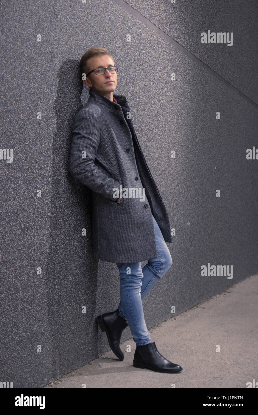 Jeune homme, 20 ans, bon, à la pose, à la forte. S'appuyant sur un mur gris, manteau gris, jeans, des bottes. Banque D'Images