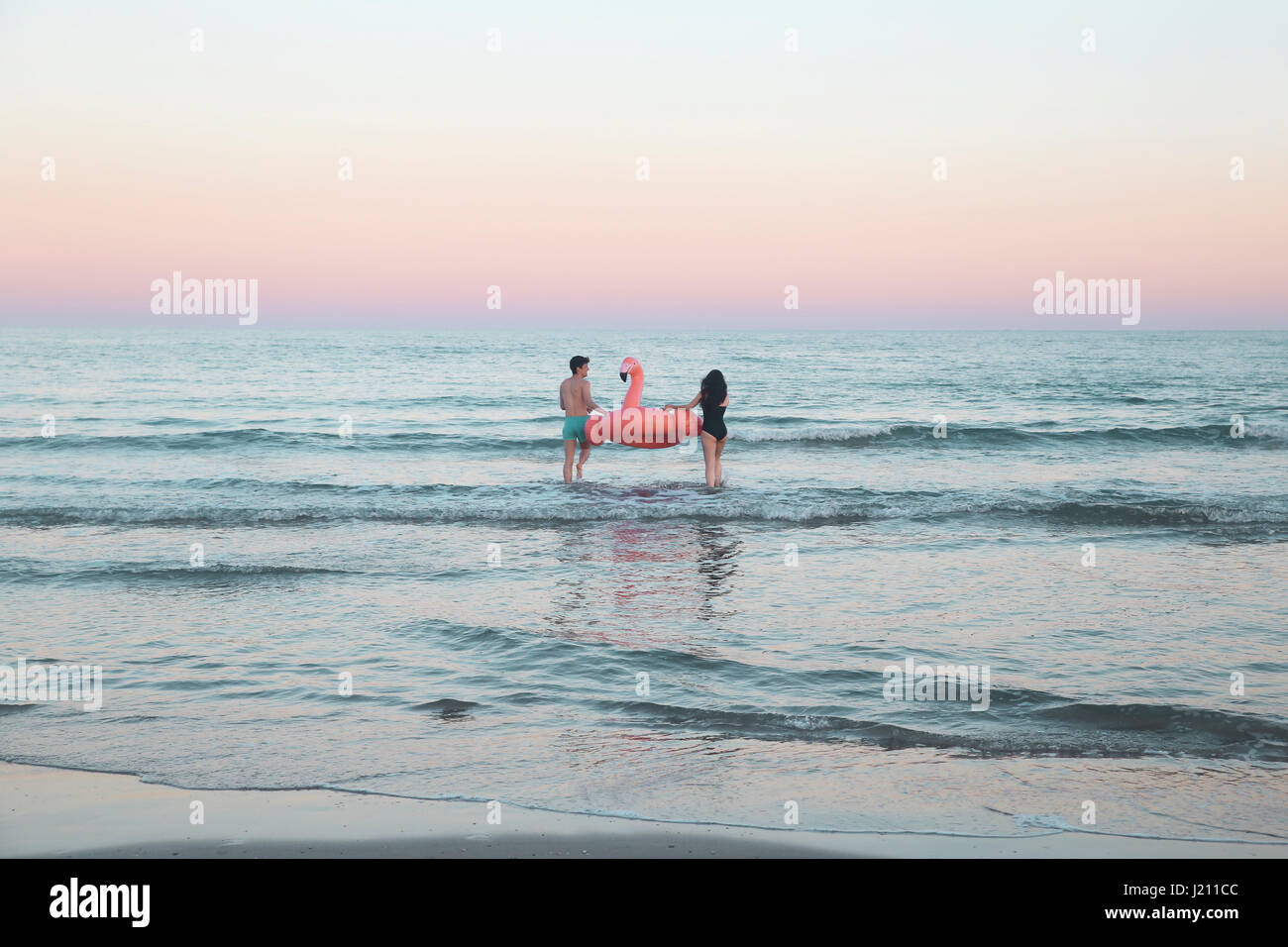 Vue arrière du jeune couple en train de marcher dans la mer avec flamant rose gonflable Banque D'Images