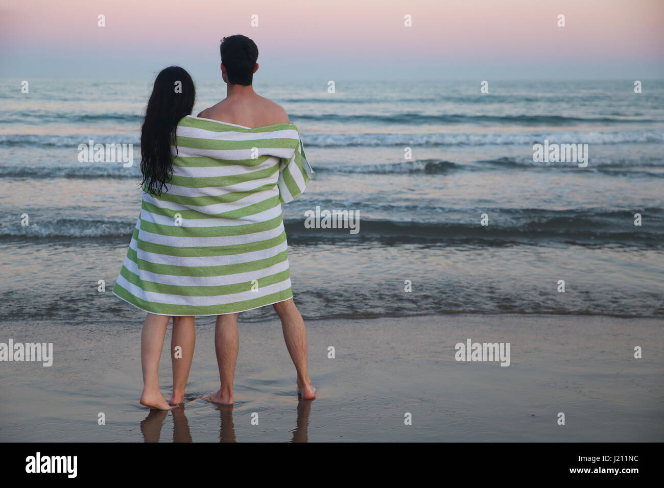 Vue arrière du jeune couple sur la plage serviette enroulée ensemble en regardant le coucher du soleil Banque D'Images