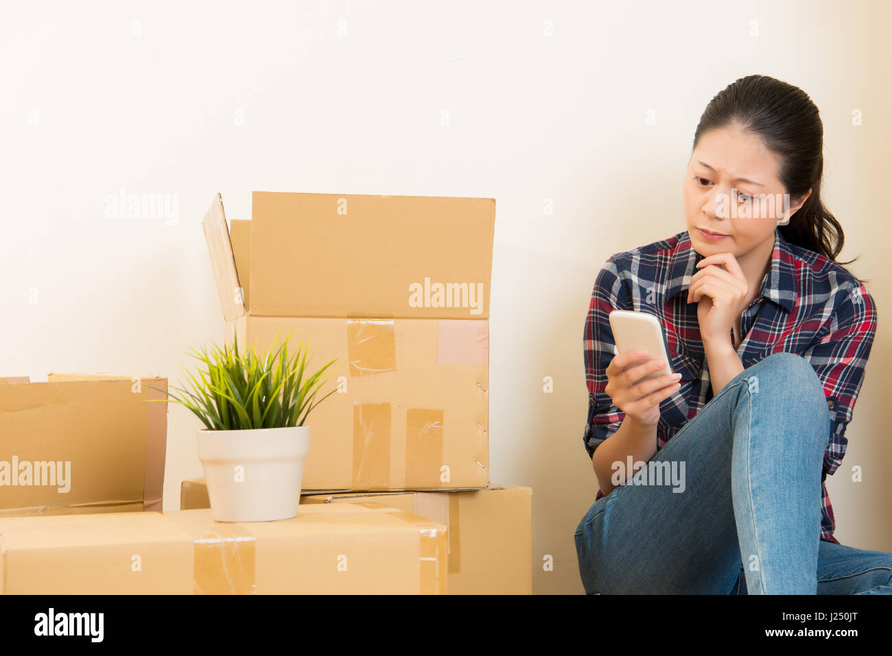 Jeune femme est un déménagement, assis parmi les boîtes de carton à l'aide d'un smartphone à la recherche de nouveaux meubles. race mixte modèle chinois asiatique. Banque D'Images