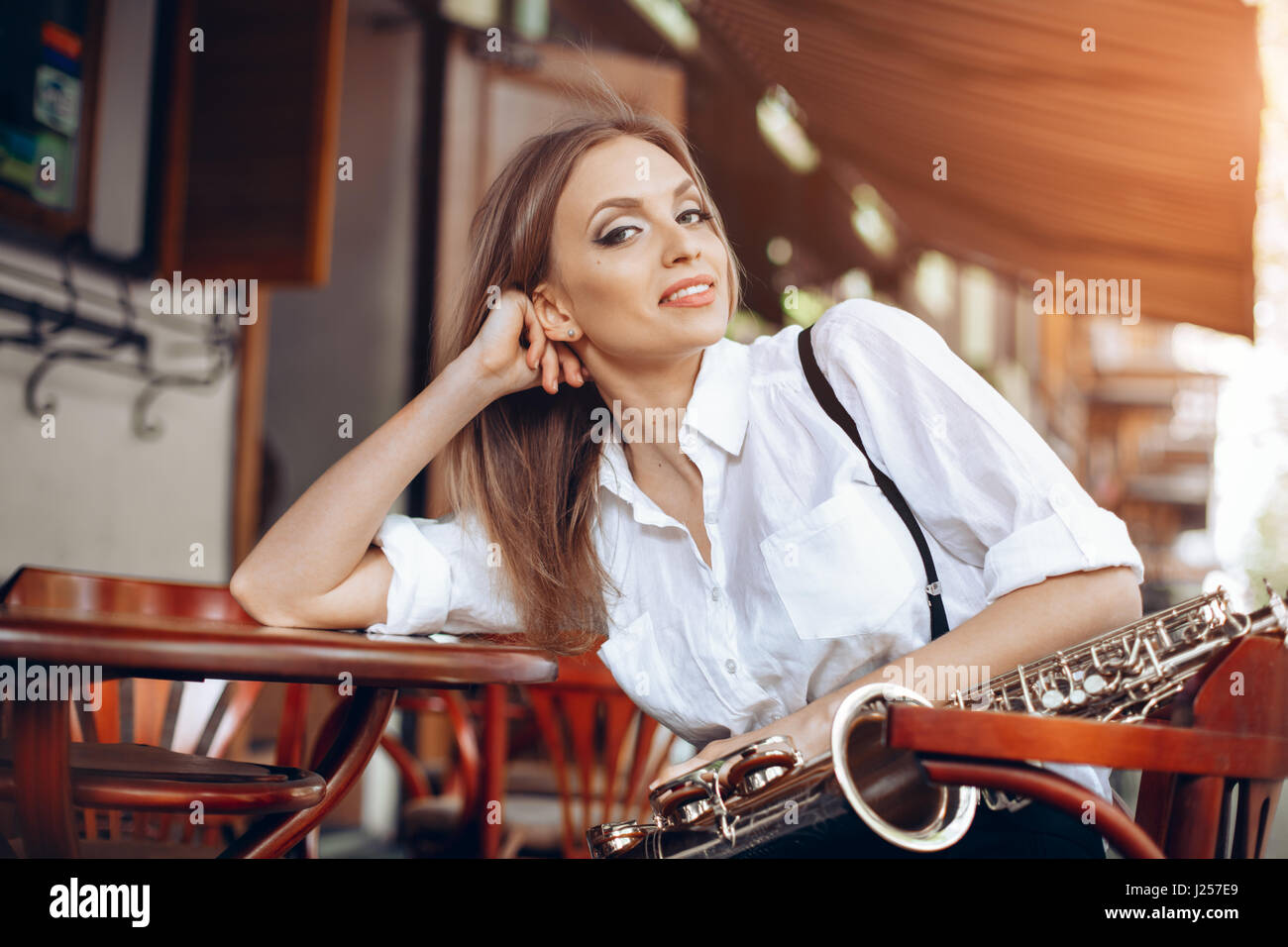 Jeune fille attirante en chemise blanche avec un saxophone assis près de caffe shop - Piscine dans sity. Sexy young woman looking at camera avec sax. Banque D'Images