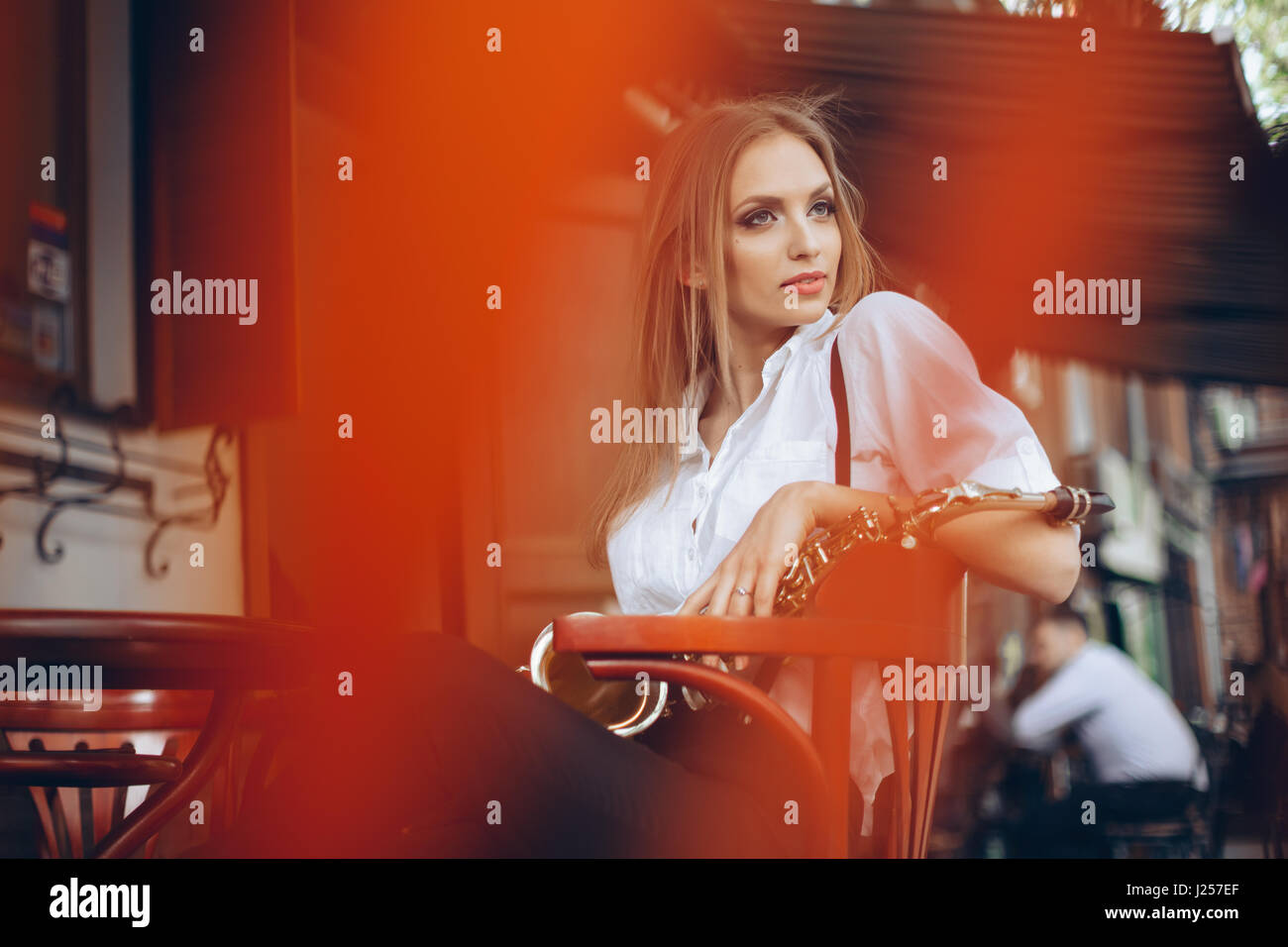 Jeune fille attirante en chemise blanche avec un saxophone assis près de caffe shop - Piscine dans sity. Jeune femme sexy avec sax pense à quelque chose. Banque D'Images