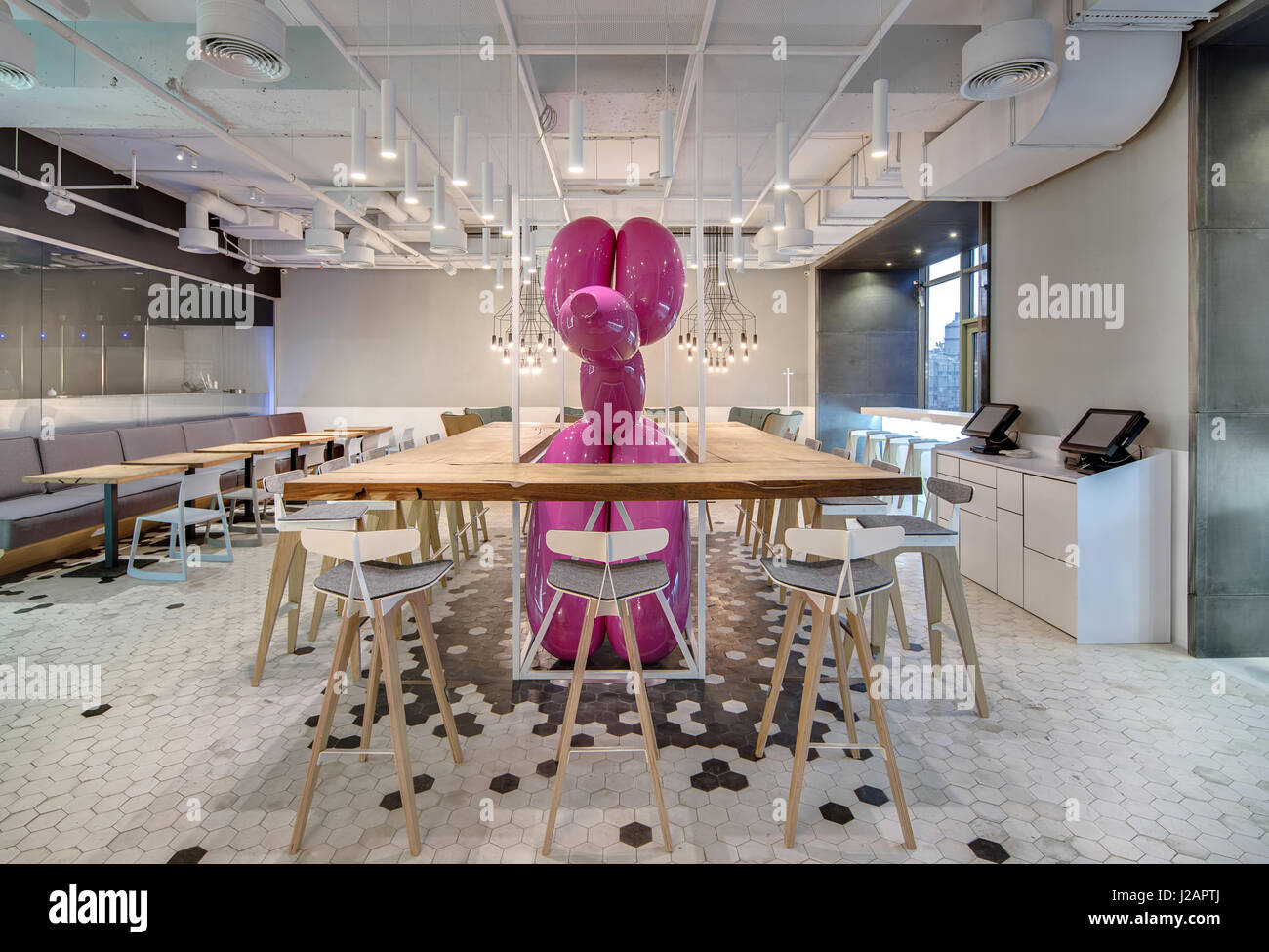 Café avec des murs clairs et carrelage noir et blanc sur le sol. Il y a des casiers en bois avec des chaises autour d'un grand chien gonflable rose, fauteuils verts, donc Banque D'Images