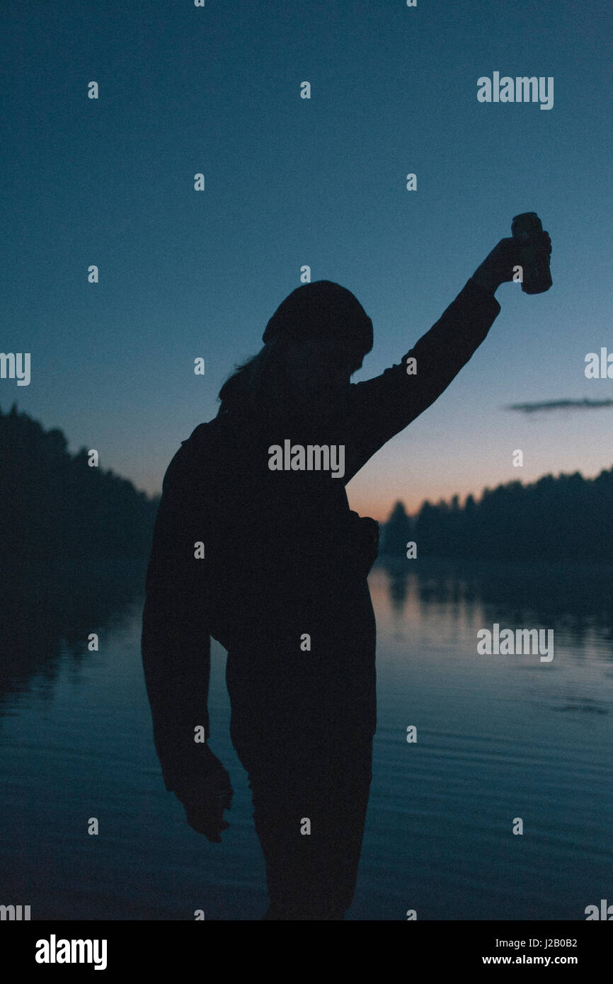 Silhouette of woman holding drink peut par le lac au coucher du soleil Banque D'Images