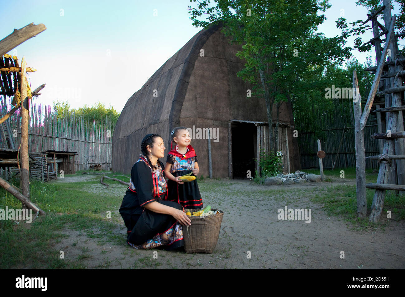 L'Ojibwe mère et fille habillé en traditionnel ruban robes avec un grand panier de maïs à l'extérieur d'une écorce couverte longue maison, lors d'une visite à l'Huron Ouendat Village indien à Midland, en Ontario. (MR) Banque D'Images