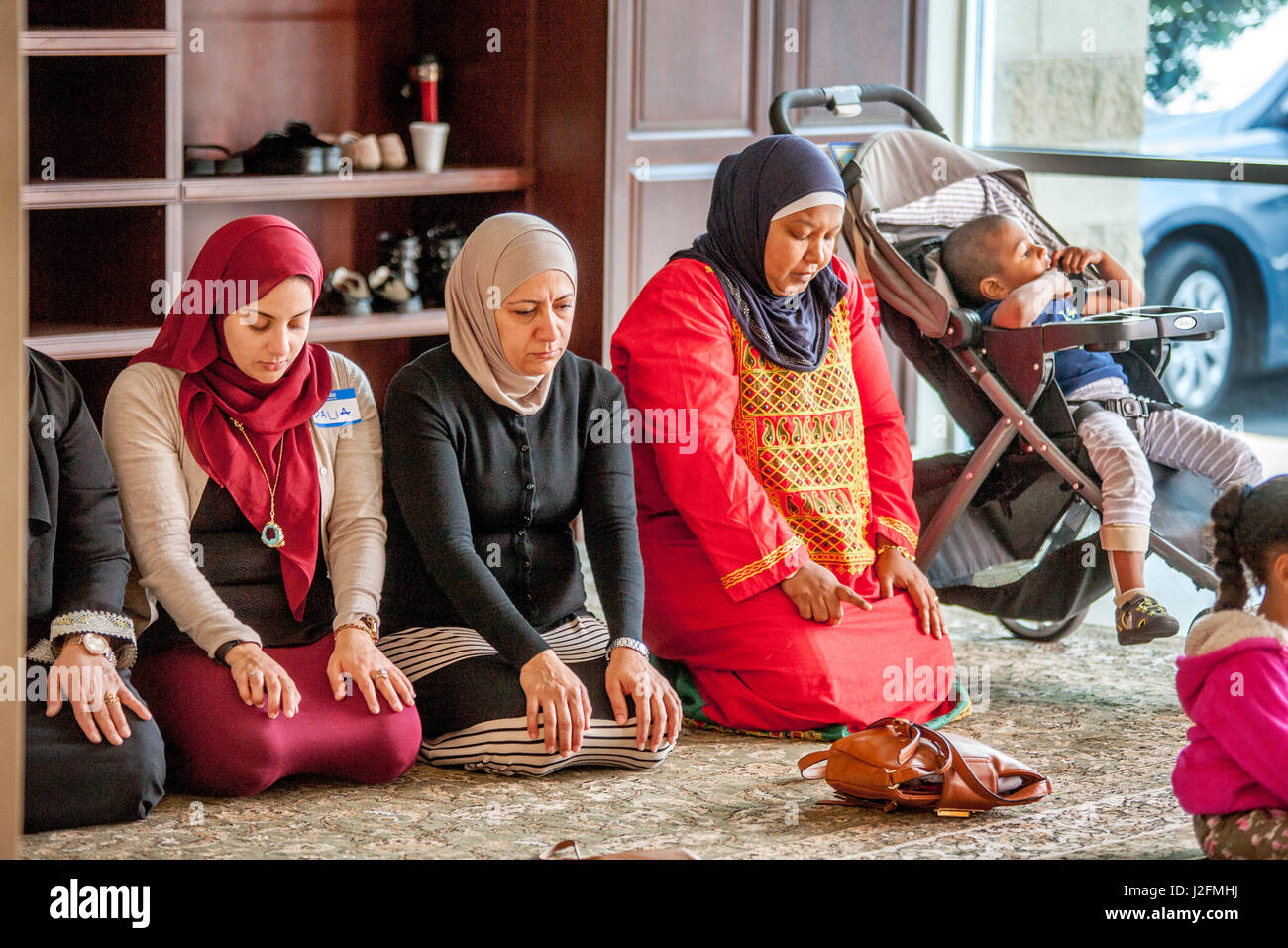 Portant un hidjab ou foulards de tête dans une pièce séparée, les femmes musulmanes s'agenouiller en prière abrahamique tandis que l'écoute de la prédication ou des services religieux pendant Khoutbah à Anaheim, CA, une mosquée. Remarque l'habillement traditionnel du Moyen-Orient et de l'enfant dans la poussette. Banque D'Images