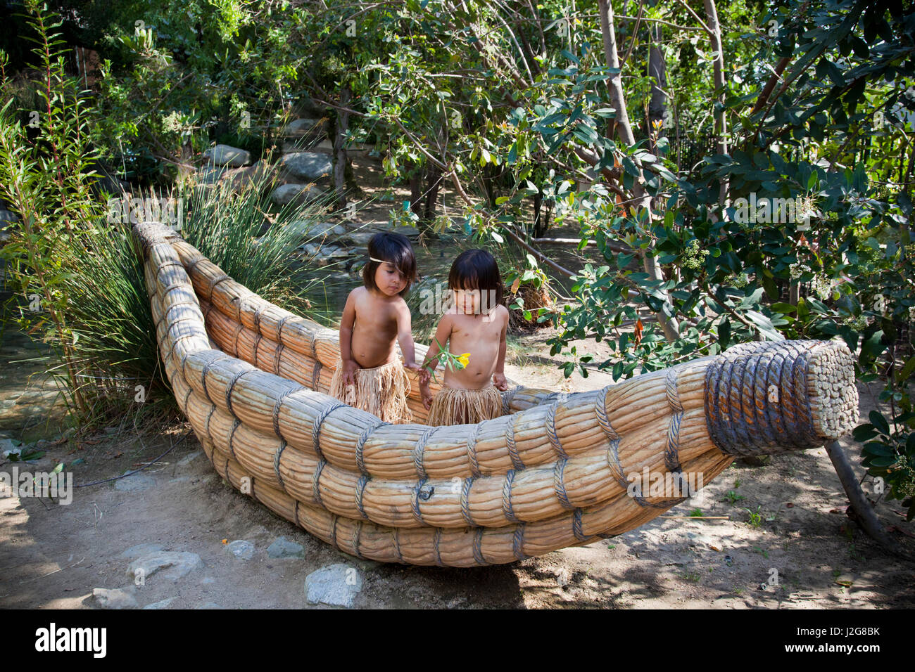 Deux California Native enfants habillés en herbe traditionnelles jupes jouer à l'intérieur d'un bateau de Tule, canot fait de queues de chat, et partager une fleur indigène. Banque D'Images