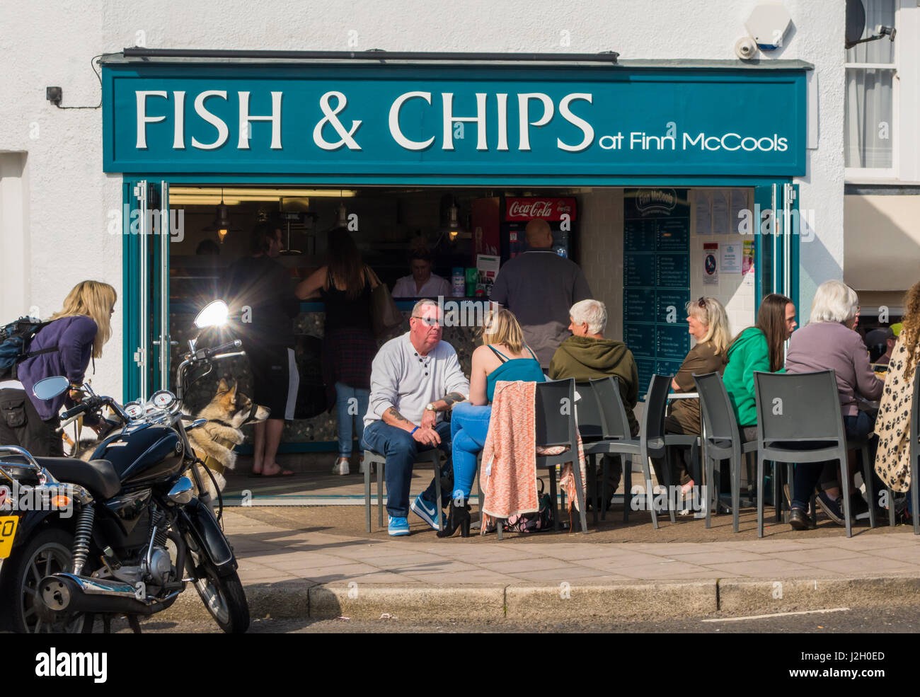 Les gens assis dehors au soleil, manger du poisson et des frites dans le cadre d'un grand panneau "fish & chips" à Teignmouth, Devon. Banque D'Images