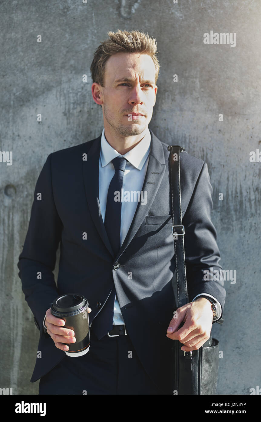 Confiant élégant businessman wearing suit avec la tasse de café en papier permanent au mur gris. Banque D'Images