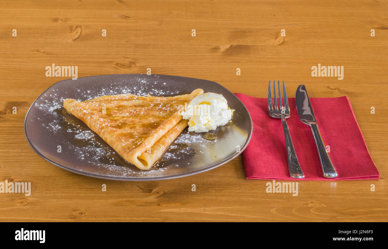 Crêpe délicieuse avec du miel et de la crème glacée servie sur une plaque gris sur une table en bois avec fourchette et couteau sur une serviette rouge Banque D'Images