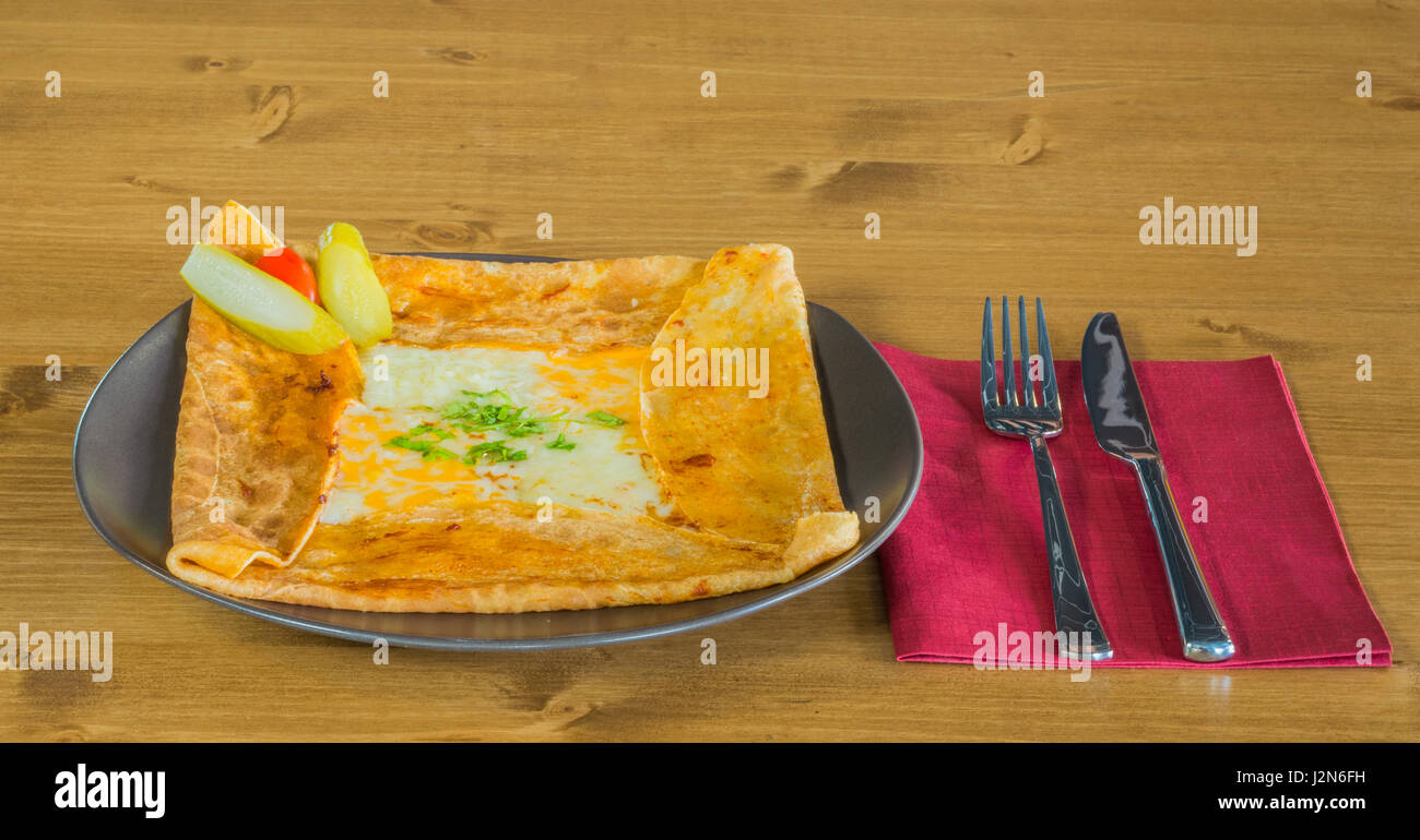 Crêpe délicieuse avec le fromage cheddar et les cornichons à côté servi sur une plaque gris sur une table en bois avec fourchette et couteau sur une serviette rouge Banque D'Images