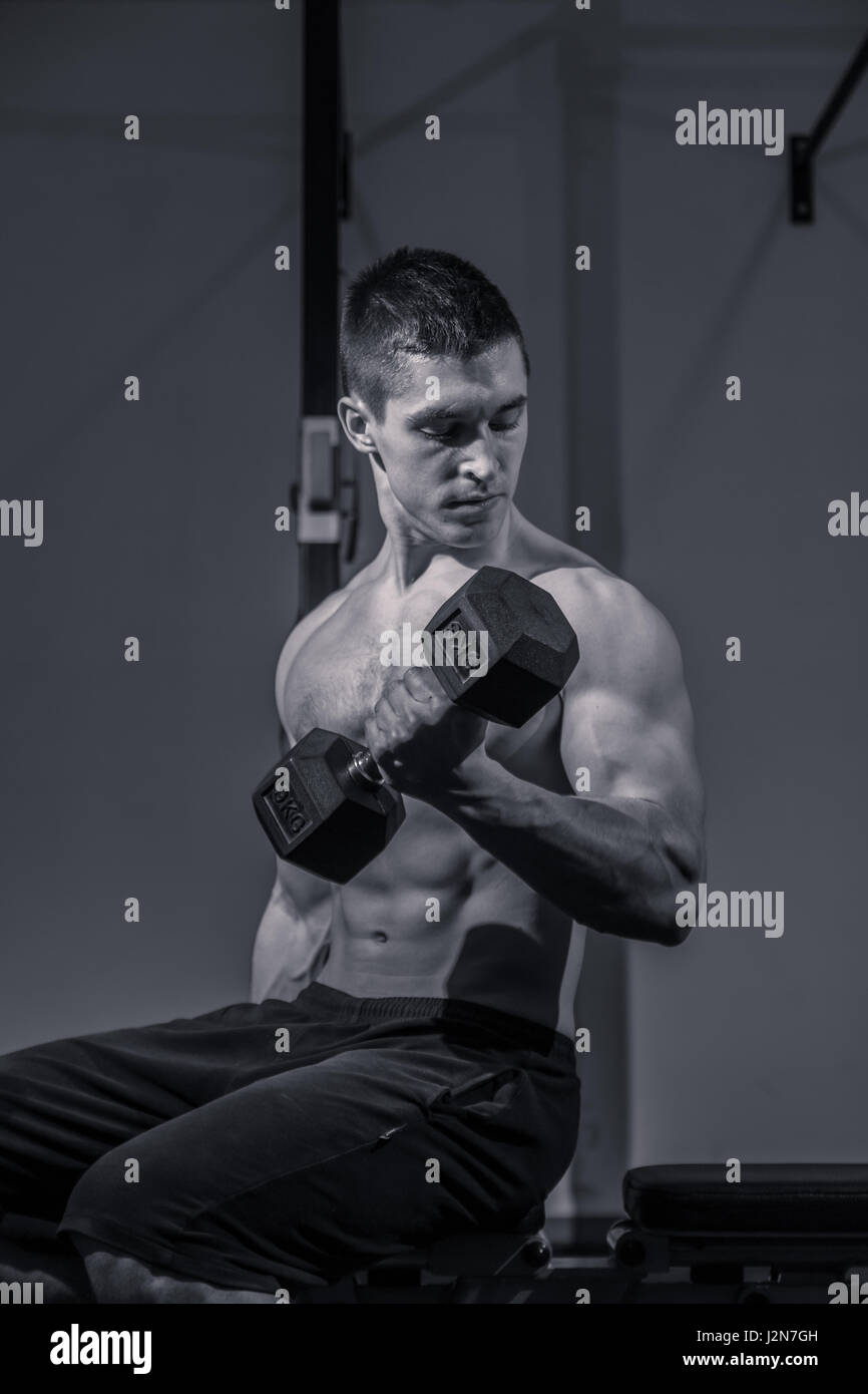 Un jeune homme, carrossier, biceps haltères d'entraînement de sport à l'intérieur sombre, noir et blanc. Banque D'Images