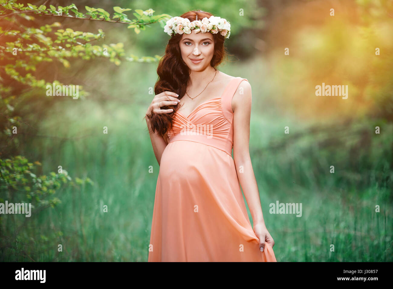 Belle jeune fille enceinte avec de longs cheveux bruns en robe de saumon avec couronne de fleurs sur la tête. Banque D'Images