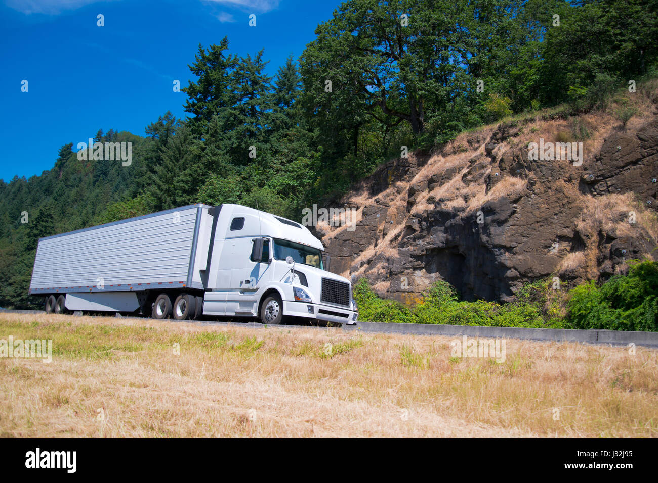 Long courrier moderne blanc et camion semi remorque frigorifique avec une unité frigorifique déménagement autoroute le long de la dernière les rochers et arbres verdoyants. Banque D'Images