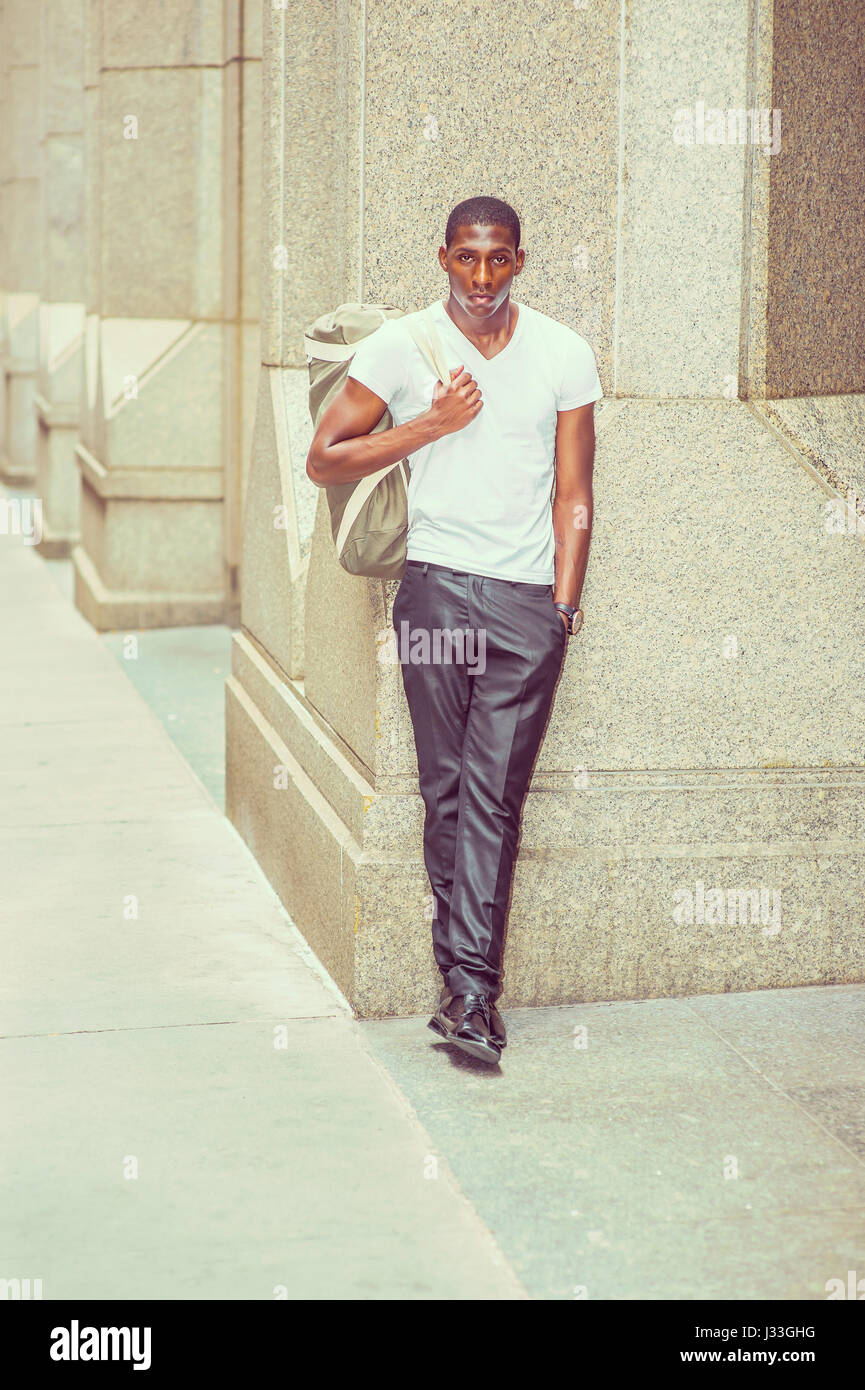 Mode de rue. Young African American Man wearing white V cou t shirt, pantalon noir, chaussures en cuir, sac de transport, à l'extérieur du bureau permanent bu Banque D'Images