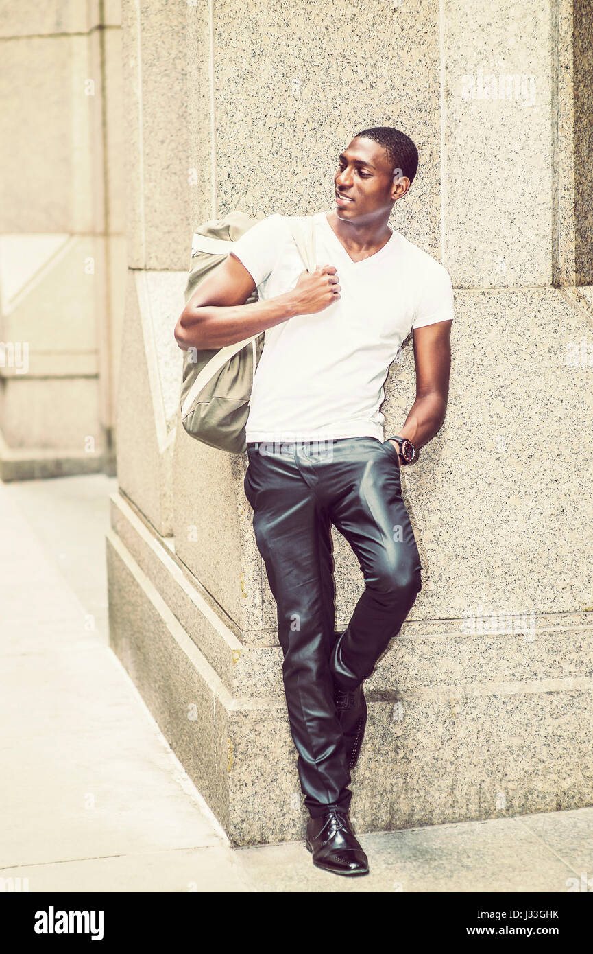 Mode de rue. Young African American man wearing white V cou t shirt, pantalon noir, chaussures en cuir, sac de transport, debout contre la colonne ou Banque D'Images