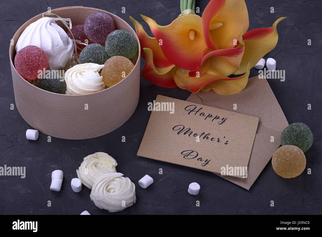 Bouquet de callas jaune rouge avec des guimauves marmelade dans une boîte ronde en bois et enveloppe avec lettrage heureux mères le background de béton gris Banque D'Images