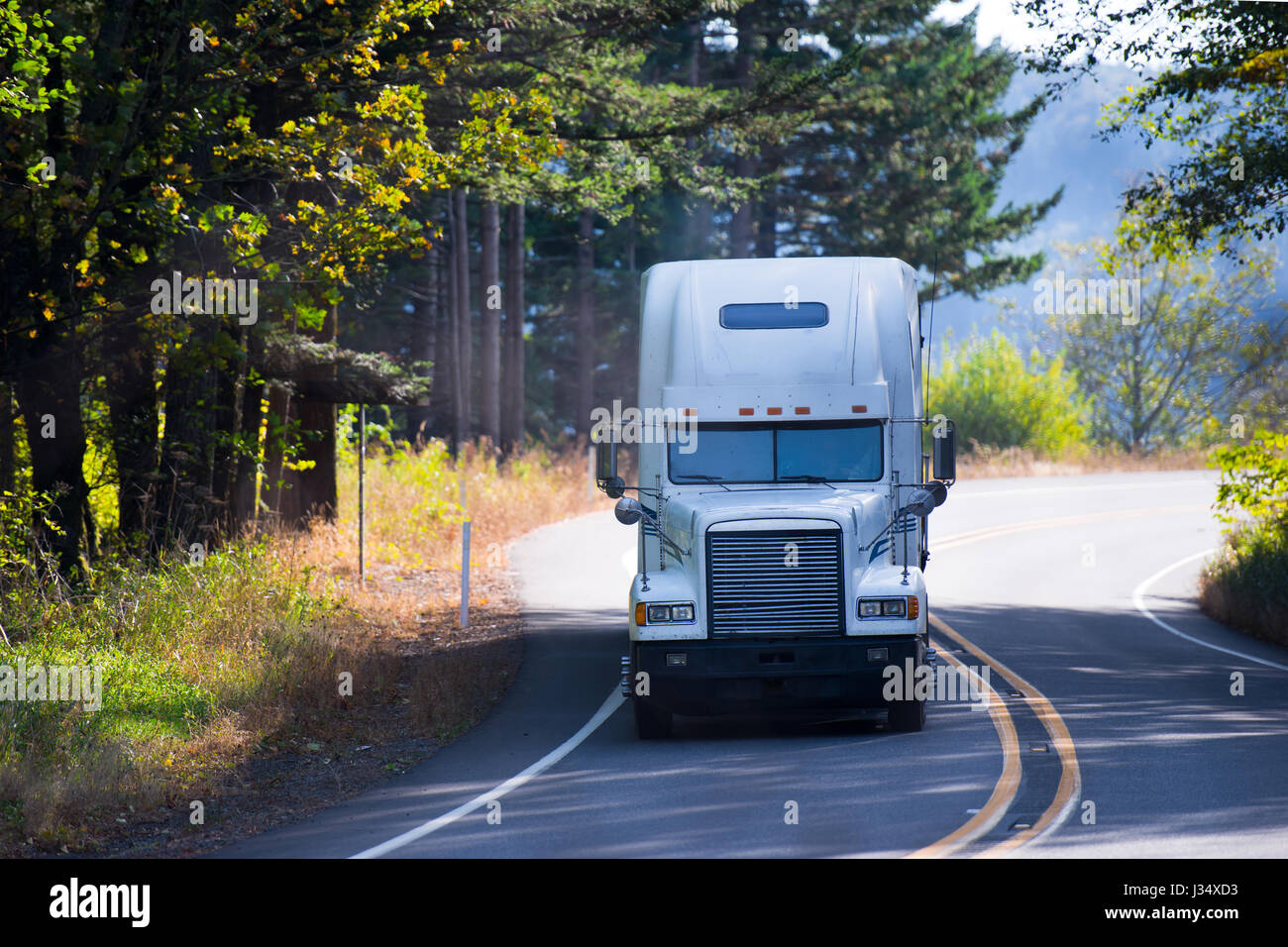 Semi-camion long-courrier classique et puissant, blanc, avec toit surélevé pour transporter des marchandises dans un virage de la route sinueuse qui traverse la forêt Banque D'Images