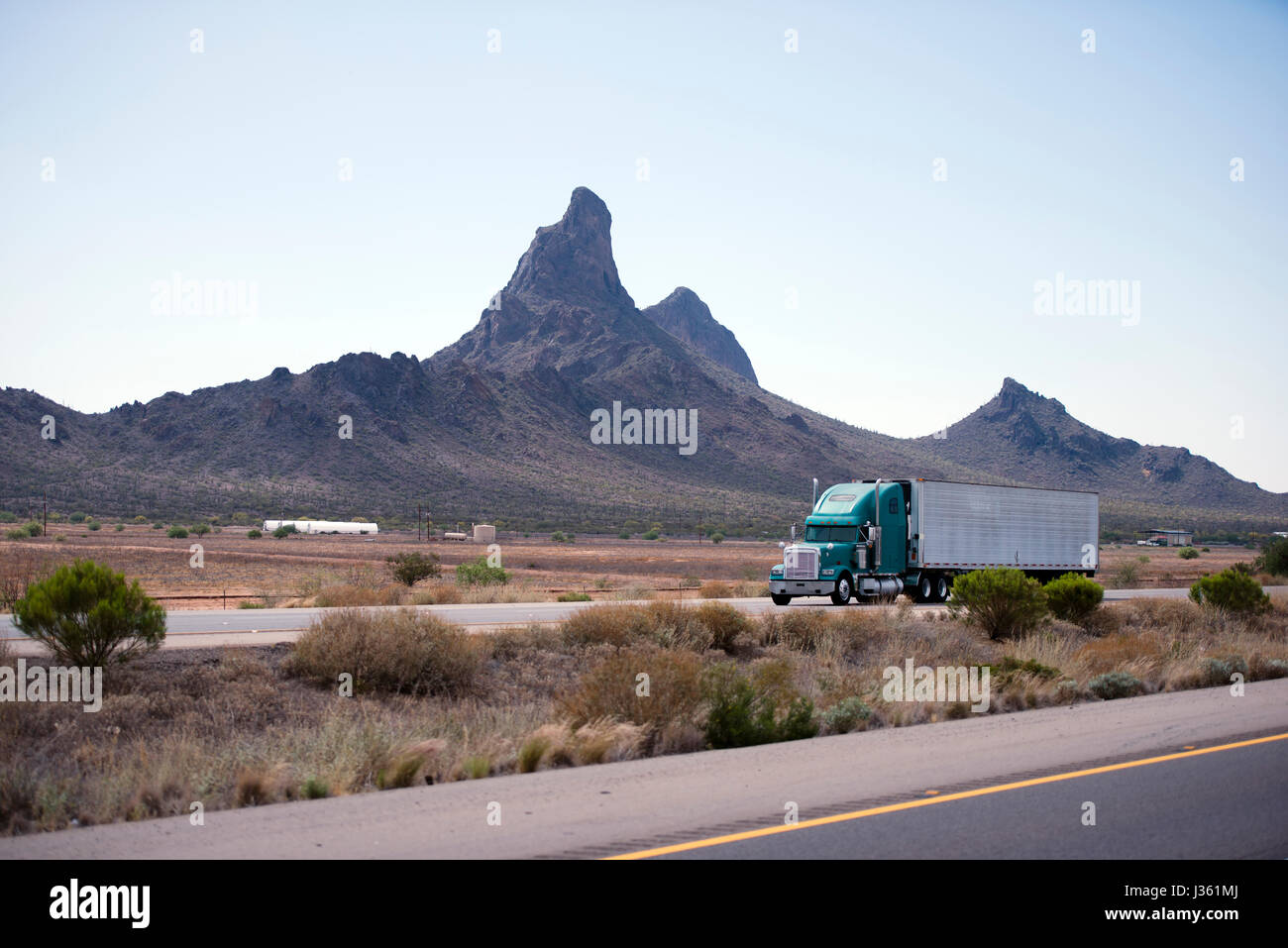 Camion vert avec un semi remorque de transport de marchandises par le routes désertes de l'Arizona avec des formes bizarres de la montagne et flétri d'arbustes. Banque D'Images