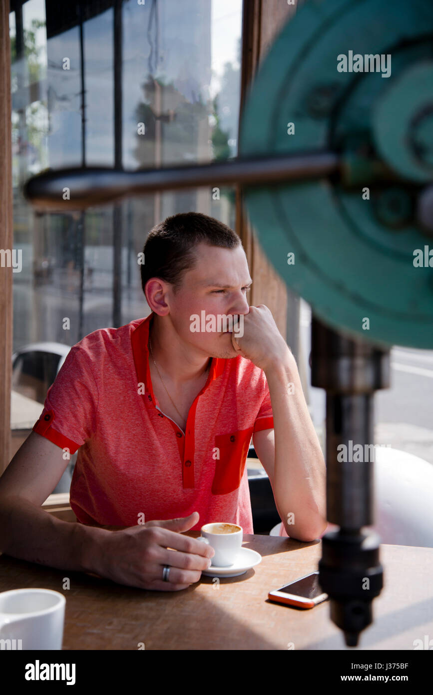 Un jeune homme aux cheveux courts à pensivement par la fenêtre tout en étant assis à une table avec une tasse de café à la main, malheureusement discuter t Banque D'Images