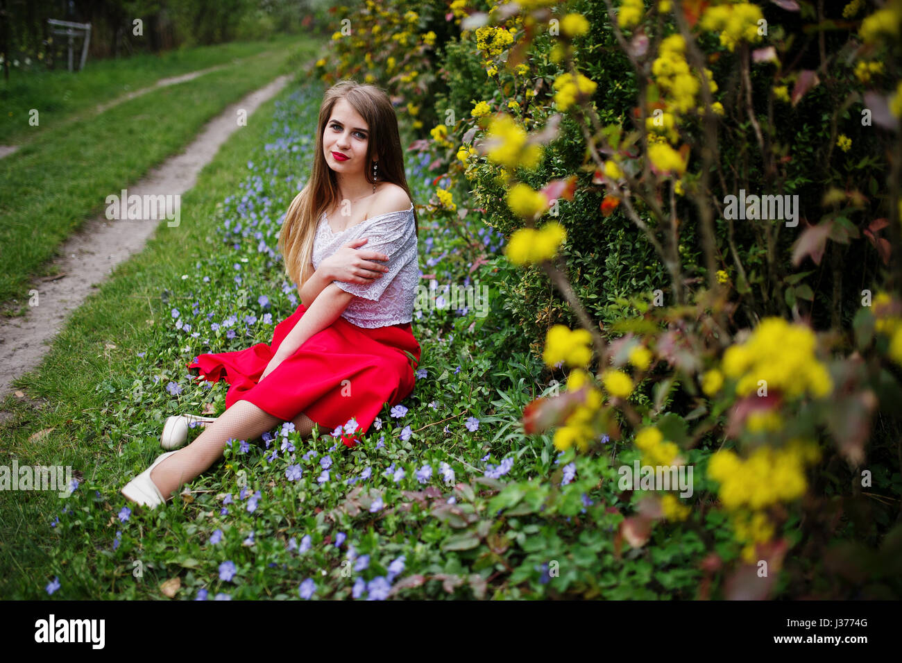 Portrait de sitiing belle fille avec lèvres rouge à fleur de printemps sur l'herbe du jardin de fleurs, l'usure de robe rouge et chemise blanche. Banque D'Images