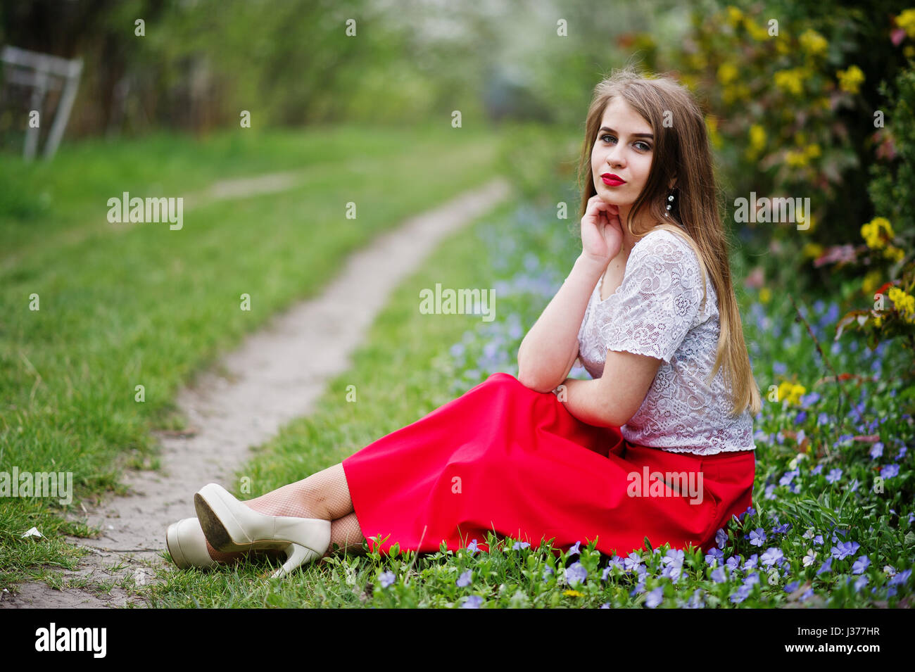 Portrait de sitiing belle fille avec lèvres rouge à fleur de printemps sur l'herbe du jardin de fleurs, l'usure de robe rouge et chemise blanche. Banque D'Images