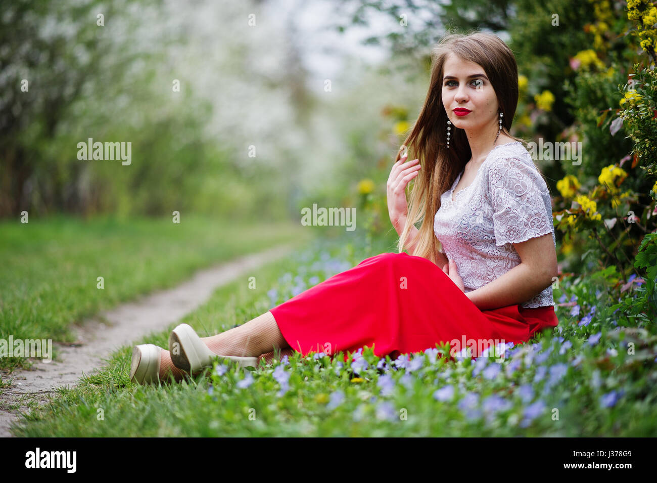 Portrait de sitiing belle fille avec lèvres rouge à fleur de printemps sur l'herbe du jardin de fleurs, l'usure de robe rouge et chemise blanche. Banque D'Images