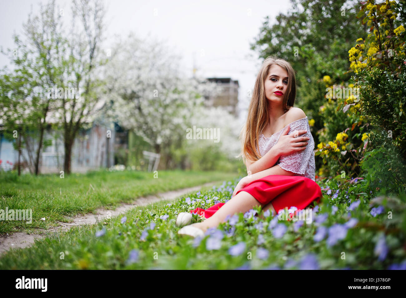 Portrait de sitiing belle fille avec lèvres rouge à fleur de printemps sur l'herbe du jardin de fleurs, l'usure de robe rouge et chemise blanche. Banque D'Images