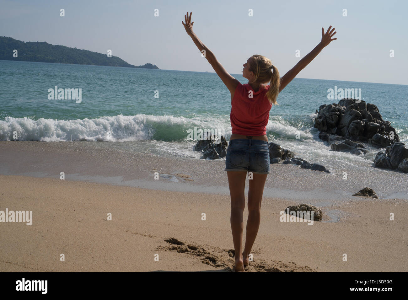 Vue arrière d'une femme en chemise rouge et bleu short debout sur la plage avec des mains et profiter de l'été sur la mer turquoise et le ciel ba Banque D'Images