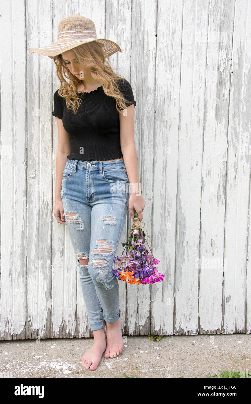 Teenage girl wearing hat et effilochée jean bleu transportant daisy bouquet par vieille grange Banque D'Images