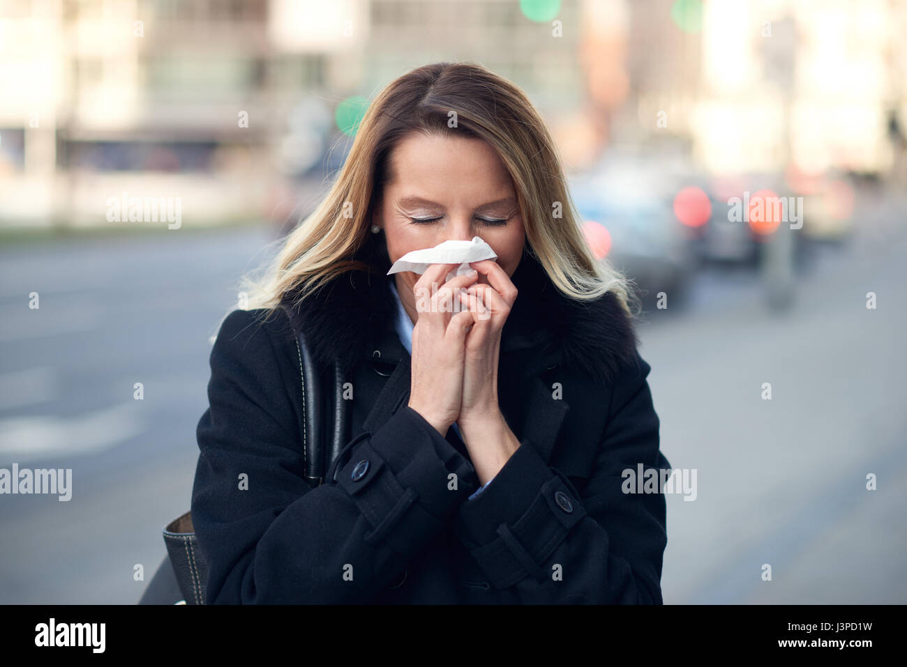 Femme avec un souffle froid hiver saison son nez sur un mouchoir ou un tissu comme elle marche dans une rue urbaine dans un concept de santé et médicaux Banque D'Images