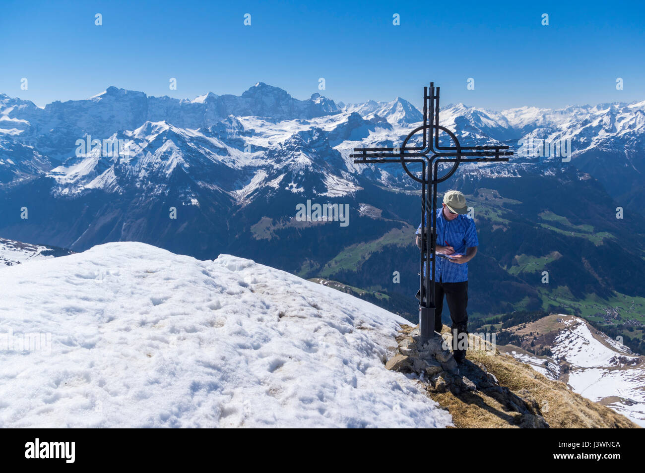 Male hiker debout à côté d'une croix au sommet du Rossstock (2461m) au printemps, écrit son nom dans le livre de sommet. Canton d'Uri, en Suisse. Banque D'Images