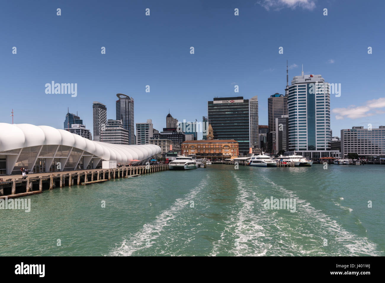 Auckland, Nouvelle-Zélande - 3 mars, 2017 : Ferry Building et pier vu de l'eau verdâtre avec des toits de la ville en arrière-plan sous ciel bleu. Ferries à doc Banque D'Images