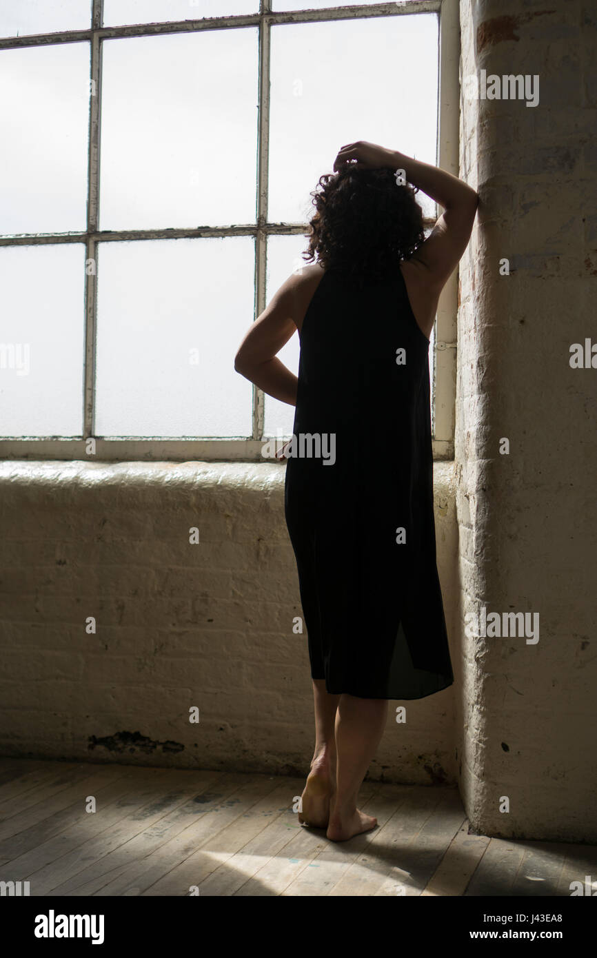 Vue arrière d'une femme portant une robe noire, de l'article par la fenêtre main sur tête appuyée contre le mur Banque D'Images