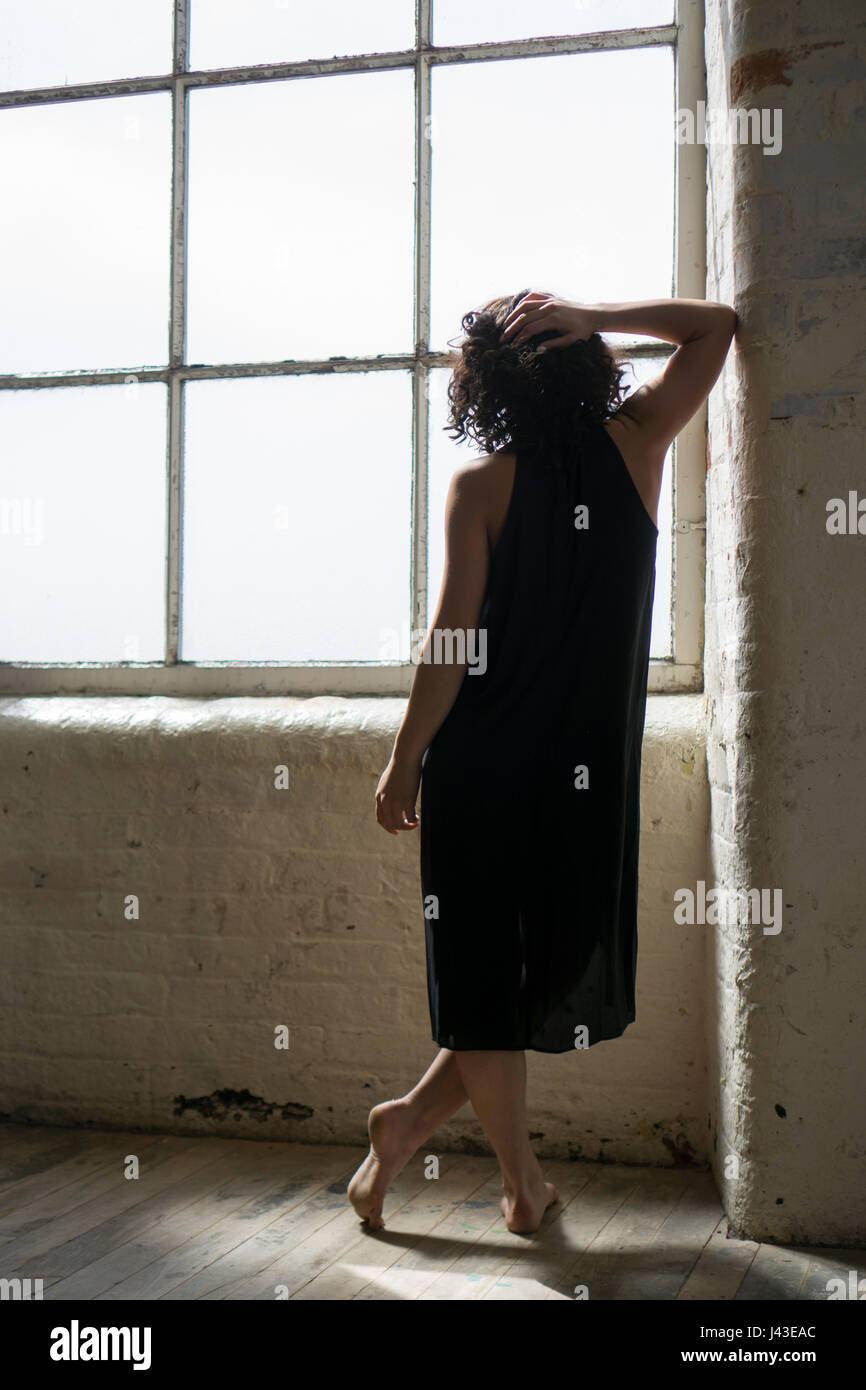 Vue arrière d'une femme portant une robe noire, de l'article par la fenêtre main sur tête appuyée contre le mur Banque D'Images