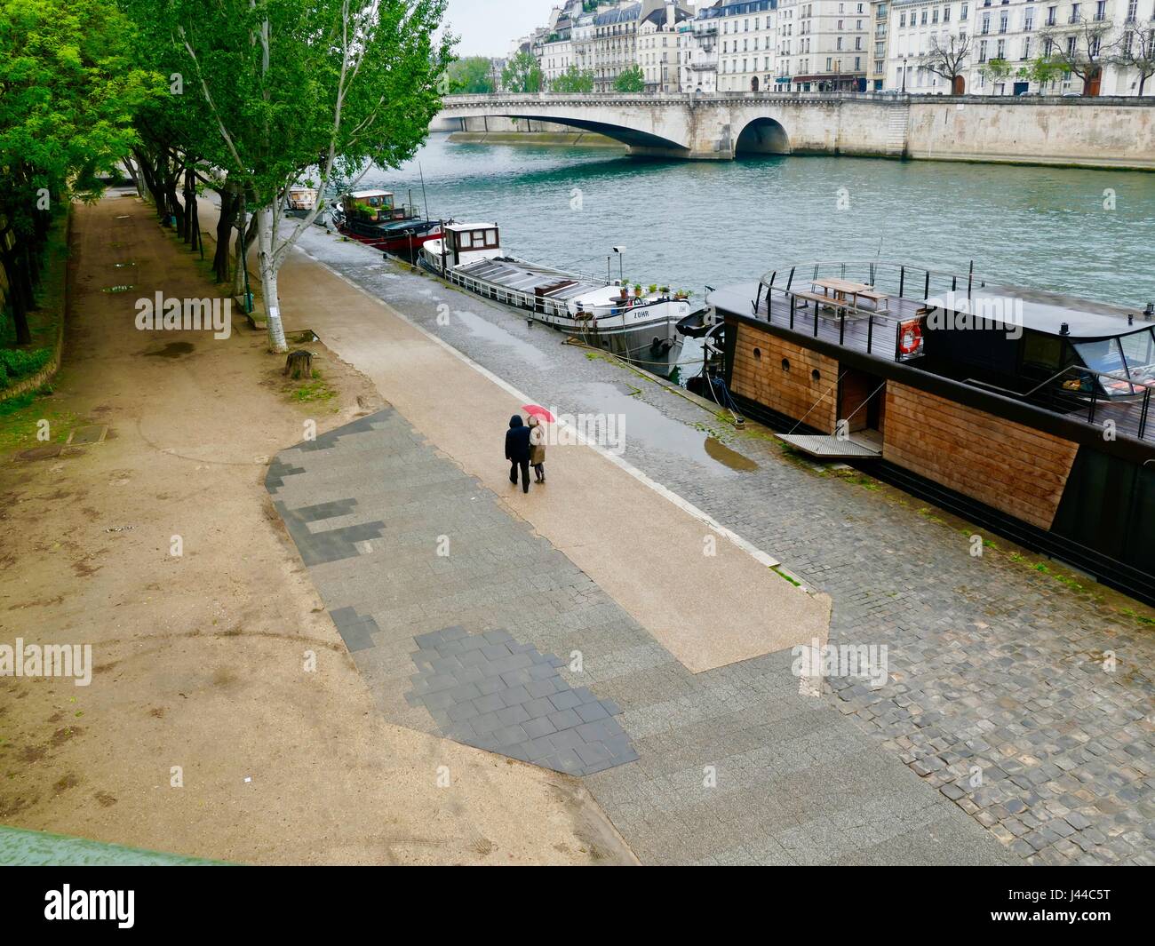 Couple d'imperméables avec parapluie rouge, marcher le long de la rivière Seine et de bateaux dans la pluie. Quai de la Tournelle, Paris, France. Banque D'Images