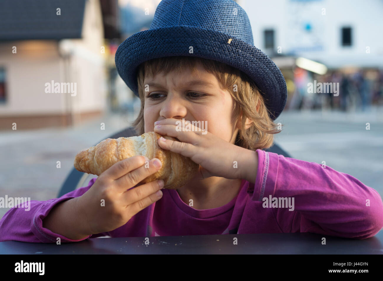 Cute little boy avec grand appétit assis à la table dévore délicieux croissant. Banque D'Images