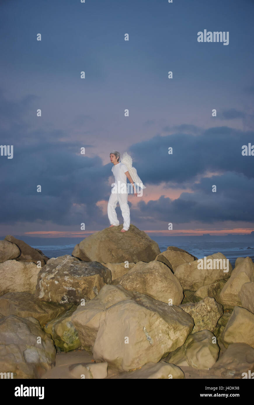 Jeune homme vêtu de blanc avec des ailes d'anges sur les rochers au bord de la mer en pleine tempête Banque D'Images