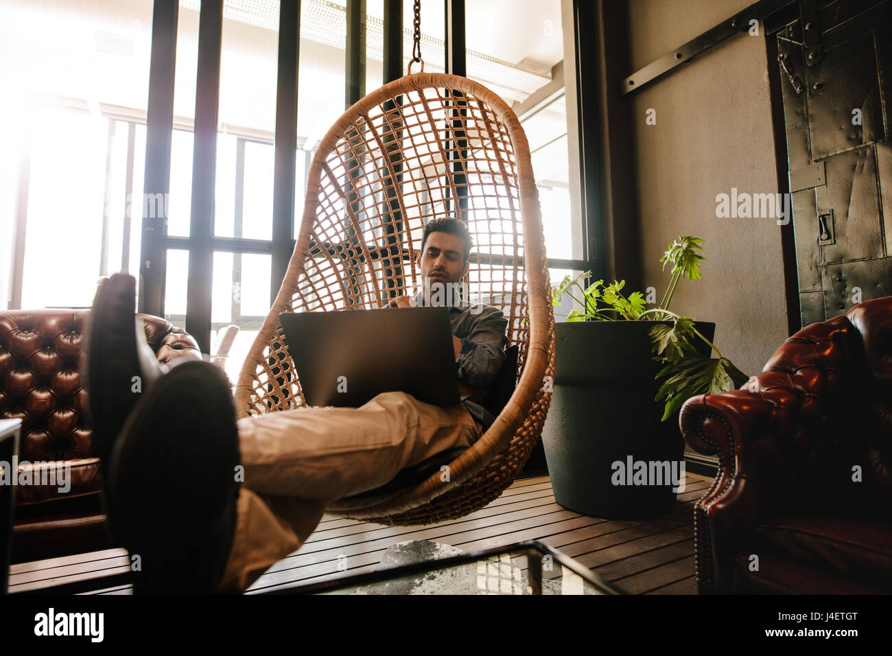 Male executive assis sur un fauteuil suspendu en osier dans office lounge and looking at laptop. Man relaxing in office lounge travaillant sur ordinateur portable. Banque D'Images