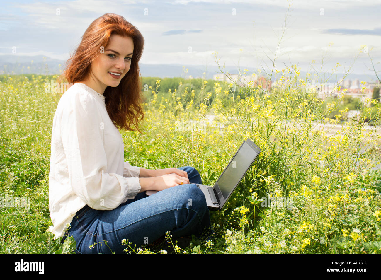 Jeune femme rousse réussie blogger sittingon l'herbe, l'écriture d'un nouveau poste pour son blog ,à l'aide d'ordinateur portable. City scape. Banque D'Images