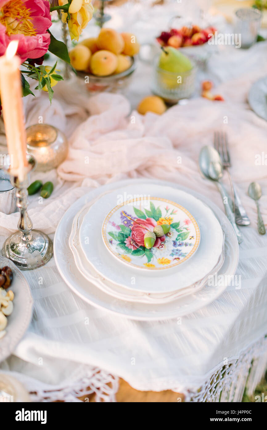 Pique-nique, nourriture, maison de vacances - maison de vacances table décoration concept avec nappe blanche, pile de plaques blanches avec des motifs floraux, deux glands sur haut, bougie allumée en chandelier, fleurs, plaques avec fruits Banque D'Images