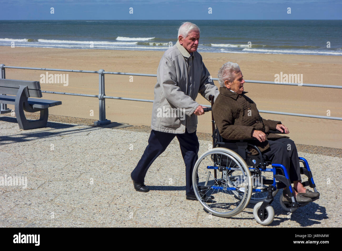 Mari à la retraite prendre femme âgées handicapées en fauteuil roulant pour une promenade sur la promenade le long de la côte par une froide journée ensoleillée au printemps Banque D'Images