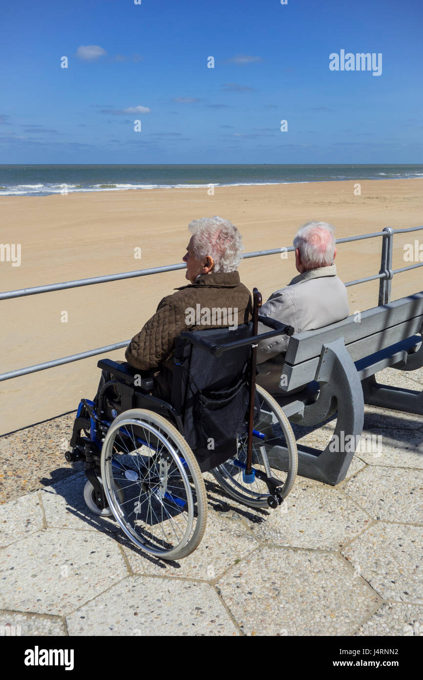 Personnes âgées handicapées woman in wheelchair et retraités mari assis sur un banc à la promenade le long de la côte par une froide journée ensoleillée au printemps Banque D'Images