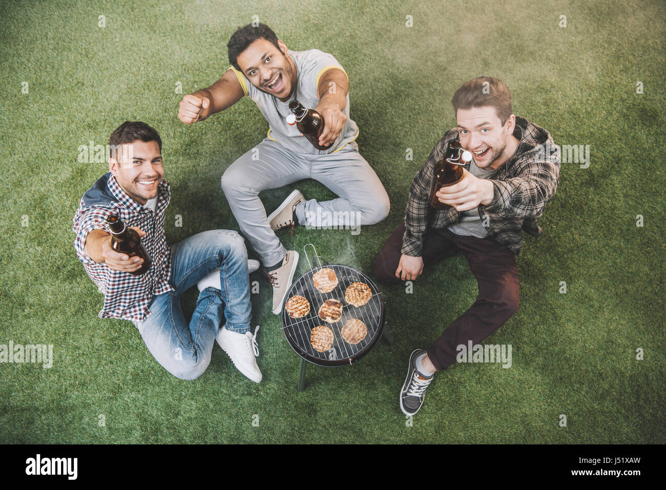 Portrait of happy young friends sitting on grass et boire de la bière pendant la cuisson de la viande Banque D'Images