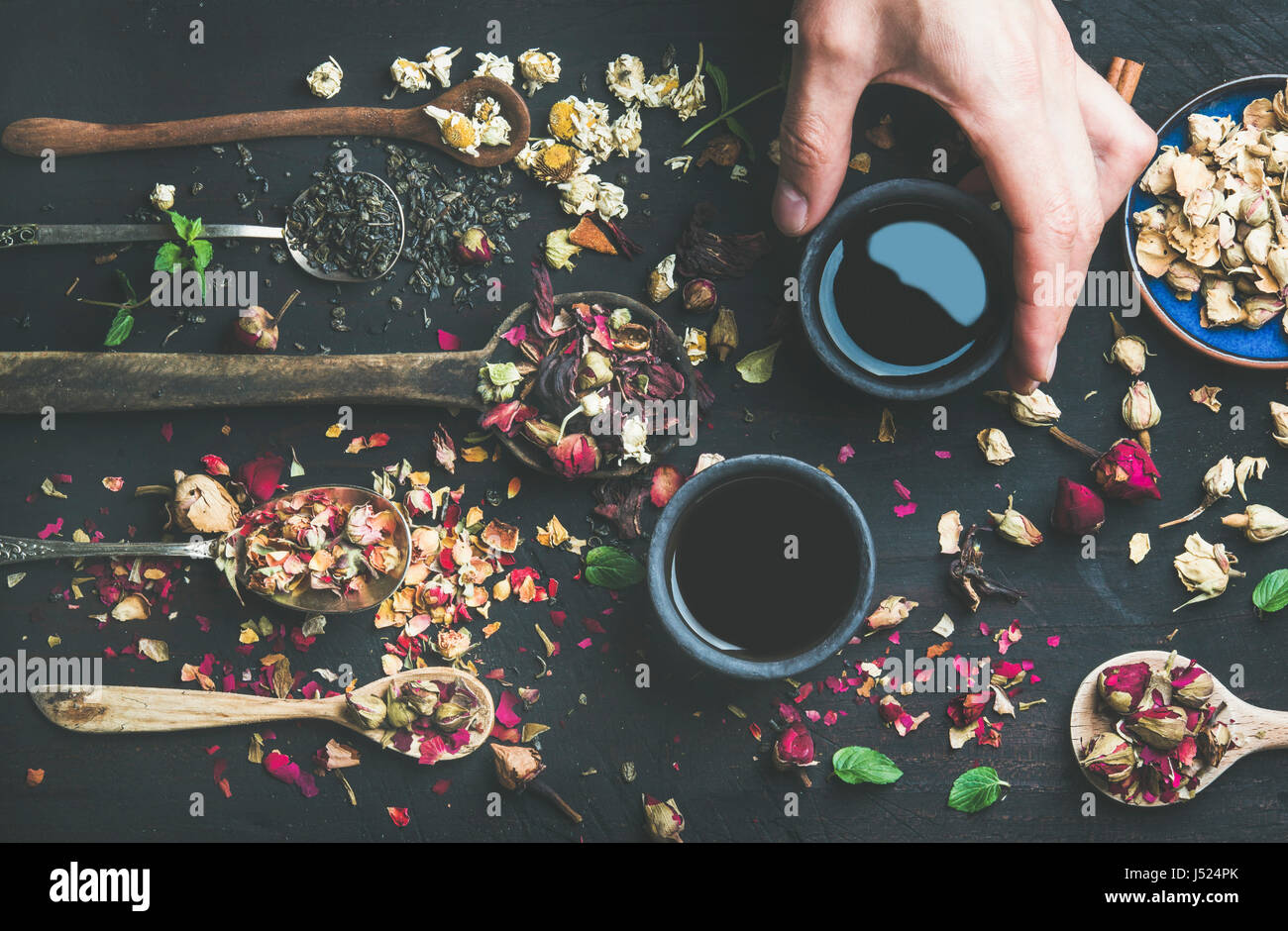 Thé noir chinois dans des tasses grès noir, la main de l'homme tenant une tasse et des cuillères en bois avec des herbes, des boutons de fleurs et feuilles sur un parquet en bois noir backgroun Banque D'Images