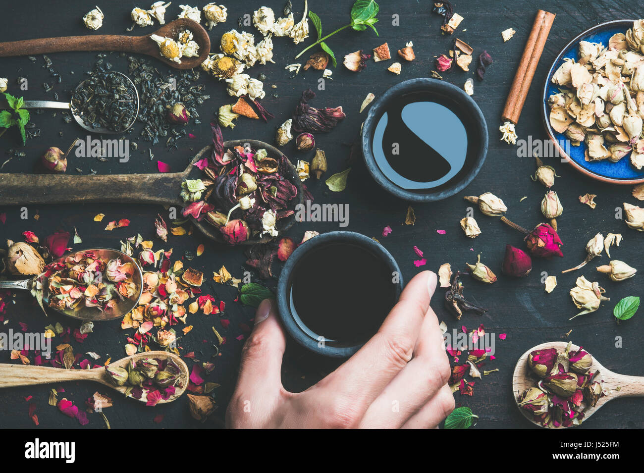 Thé noir chinois dans des tasses grès noir, la main de l'homme tenant une tasse et des cuillères en bois avec des herbes sèches, des boutons de fleurs et feuilles sur un parquet en bois noir backg Banque D'Images